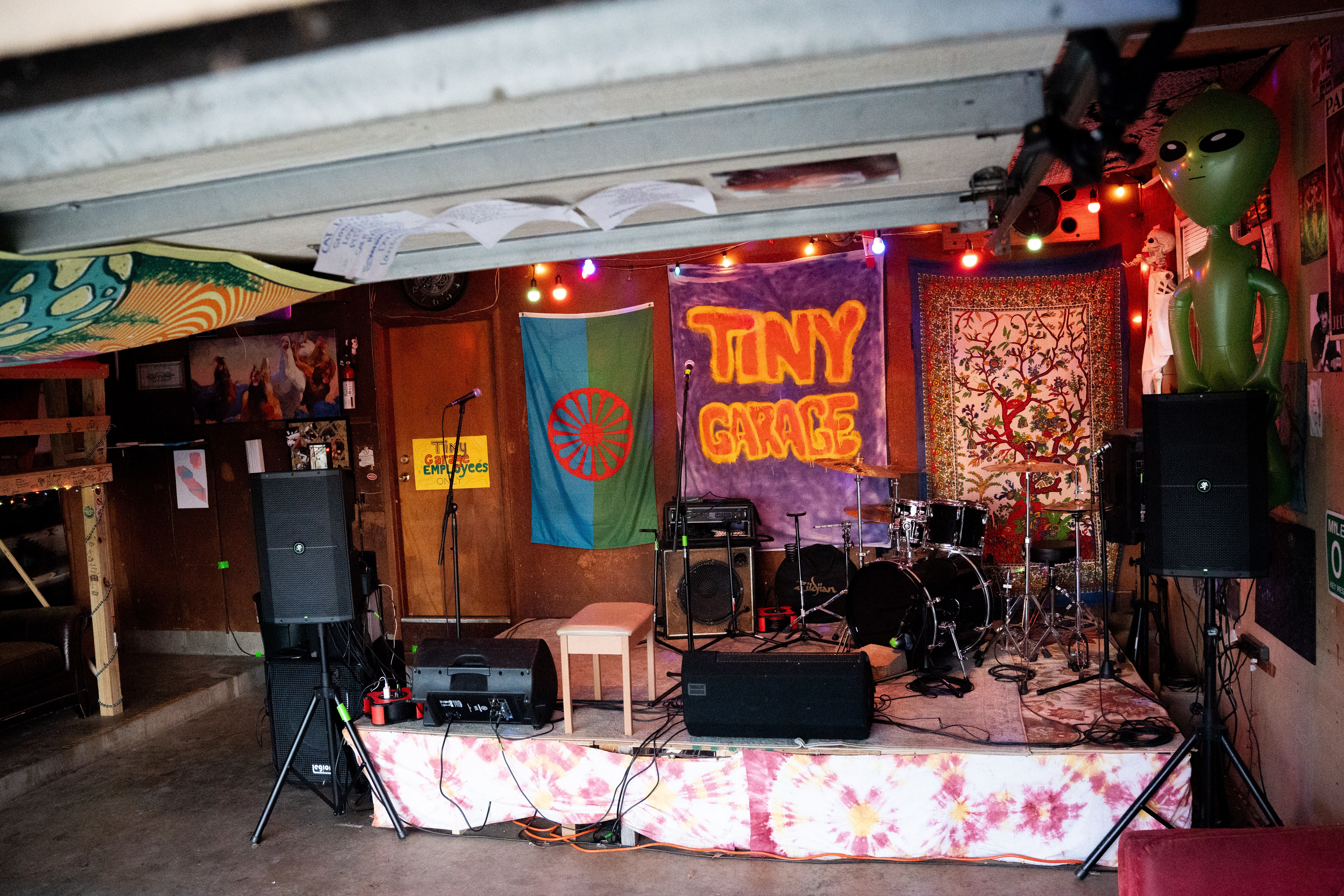 An empty stage within a garage with musical equipment sitting on it. Among other decoration is a 'Tiny Garage' banner on the back wall.