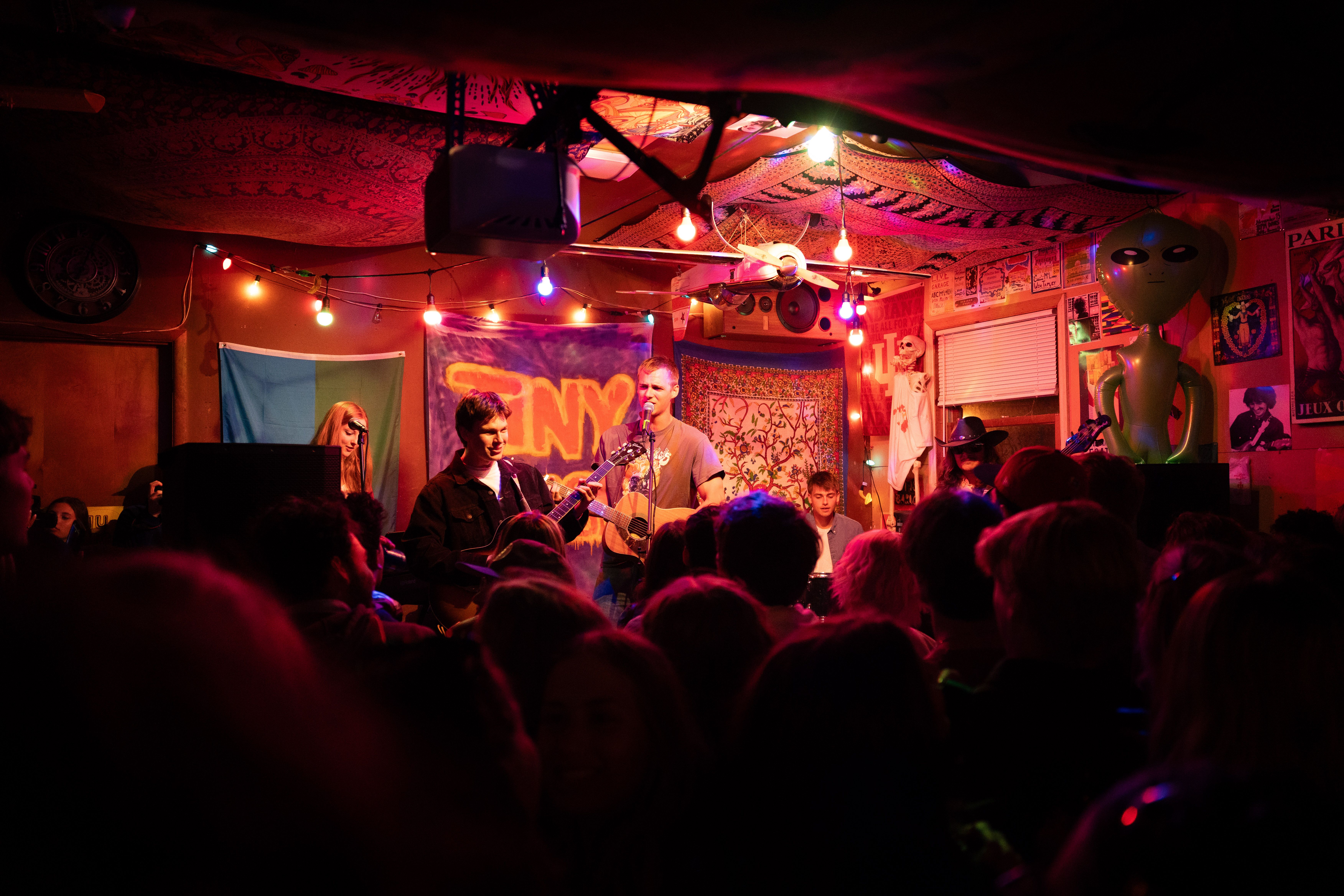 Band on stage in the back of a garage, with a crowd facing away from camera in the foreground.